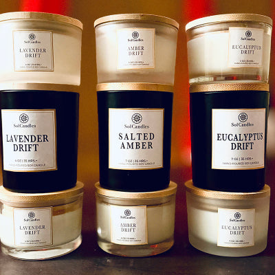 Three candles with labels indicating scents and brand name on a reflective surface.